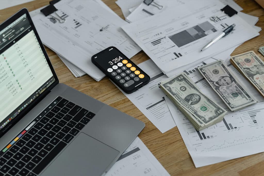 Laptop, cash, and financial documents on a desk showing budget planning related to electrician salary.